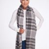 An older woman with short gray hair smiles warmly while wearing the Woolx Bradie Scarf in dark grey plaid, paired with a white long-sleeve top and blue jeans. She exudes confidence as she stands against a simple white background.