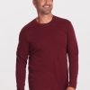 A man wearing a Woolx Easton Crew Neck Shirt in Cranberry Melange and blue jeans is smiling while standing against a plain white background.