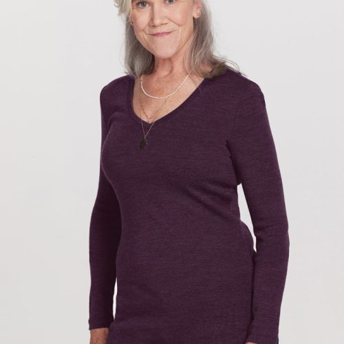 An older woman with gray hair stands against a plain background, wearing the Woolx Eva Tunic - Deep Plum and jeans. The long-sleeved tunic is made of odor-resistant Merino wool, adding to her comfort. She has a gentle smile and is accessorized with a necklace.