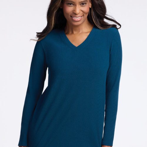 A woman is smiling, wrapped in a cozy Evie Tunic by Woolx, a long-sleeved merino wool sweater in Real Teal with a v-neck design. Her long hair cascades freely as she stands against a plain white background. | Keesha is 5’9.5”, wearing a size S