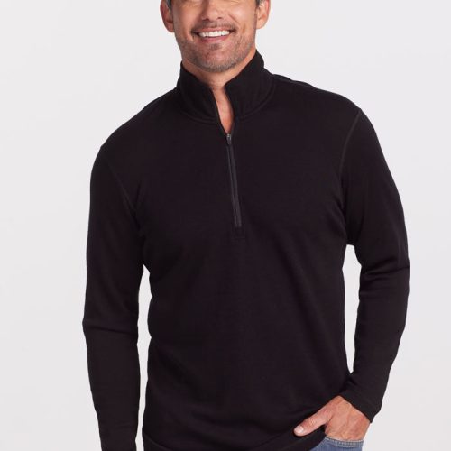 A person with short, gray hair is smiling and standing against a plain white background. They are wearing an ultra-soft Explorer 1/4 Zip pullover in black from Woolx, made of midweight fabric, paired with blue jeans. One hand is in their pocket while the other rests at their side.