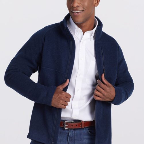 A person wearing a dark navy Fairbanks Jacket from Woolx over a white button-up shirt and blue jeans poses confidently. They are smiling and adjusting their ultra-heavyweight jacket with both hands against a plain, light-colored background. | Trell is 6’2”, wearing a size M