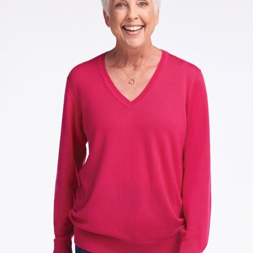 A smiling older woman with short white hair is standing against a plain background. She is wearing the Francesca Sweater - Berry Burst by Woolx and blue jeans. | Kathy is 5'9", wearing a size S