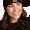 A woman with long brown hair smiles while wearing the Woolx Baylor Beanie in French Roast, distinguished by its brown patch. Her black sweater complements the stretchy foldable ear cuff as she poses with one hand adjusting her hat against a plain white background.