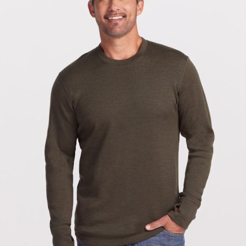 A person with short dark hair and a beard is smiling while wearing the Glacier Base Layer Top in Bark by Woolx and blue jeans. They stand with one hand in their pocket against a plain white background, exuding comfort and style.