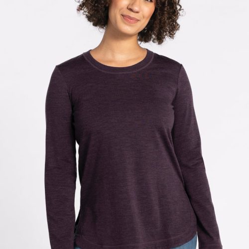 A person with curly hair is smiling while wearing a relaxed-fit, long-sleeved Hazel Tunic in Deep Plum from Woolx and blue jeans. The background is plain white. | Tori is 5'7", wearing a size S