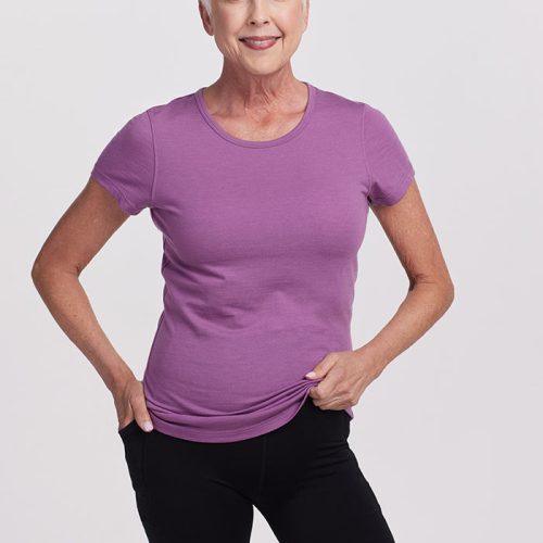 An older woman with short white hair is wearing the ultra-soft Dewberry Addie Short Sleeve Crew by Woolx. She stands smiling with one hand on her hip and the other holding the hem of her merino tee, paired with black shorts. The light-colored, plain background enhances the vibrancy of her outfit.