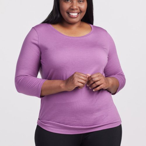 A person with long hair smiles at the camera, wearing a Woolx Jenny 3/4 Sleeve Crew Neck in Dewberry and black pants. They stand against a plain white background, exuding a sense of casual comfort.