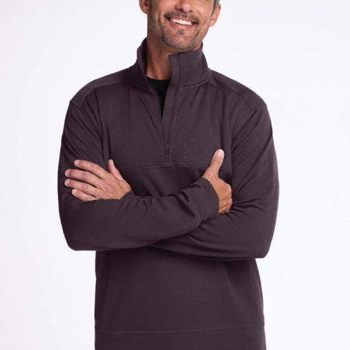 A man with short hair and a beard smiles while crossing his arms. He is wearing a dark Mason 1/4 Zip - Chocolate Plum pullover by Woolx and blue jeans, standing against a plain white background.