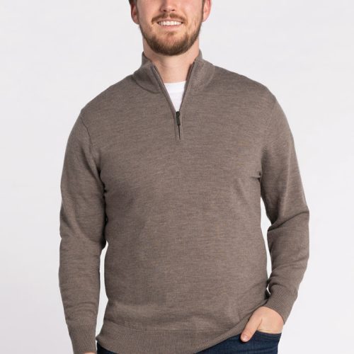 A man wearing the Enzo Quarter Zip in Sable from Woolx, paired with dark jeans, stands against a plain white background. He is smiling with one hand in his pocket. | Will is 6', wearing a size M