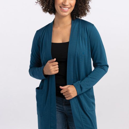 A person with curly hair smiles while wearing the Woolx Paisley Cardigan in Real Teal over a black top and blue jeans. They are standing against a plain white background, with one hand holding the edge of the cardigan made from a Duralite blend. | Tori is 5'7", wearing a size S