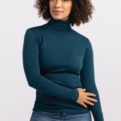 A person with curly hair poses against a plain background, wearing the Peyton Turtleneck in Real Teal from Woolx and blue jeans. They gently rest one hand on their stomach while the other arm hangs loosely by their side, with a composed expression on their face. | Tori is 5'7", wearing a size S