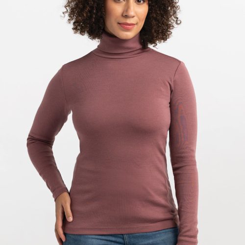 A person with curly hair wearing a Wild Ginger Peyton Turtleneck from Woolx and blue jeans stands against a plain white background. They are facing the camera with a neutral expression and one hand on their hip, ready for any adventure. | Tori is 5'7", wearing a size S