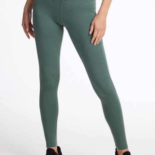 A person's lower body is visible against a white background, wearing Duck Green Piper Pocket Leggings by Woolx paired with black sneakers.