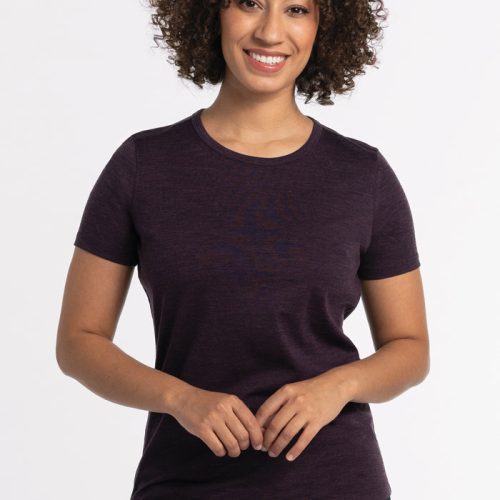 A person with curly hair wearing the Woolx Liza Crew Neck Tee in Deep Plum and blue jeans stands against a white background, smiling. They have their hands clasped in front of them. | Tori is 5'7", wearing a size S