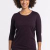 A person with curly hair smiles while standing against a white background, wearing the Woolx Jenny 3/4 Sleeve Crew Neck in Deep Plum paired with dark jeans. | Tori is 5'7", wearing a size S