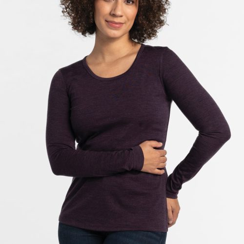 A person with curly hair is wearing the Remi Long Sleeve T-Shirt in Deep Plum by Woolx and a pair of jeans. They stand against a plain white background, smiling gently with their arms relaxed, enjoying the comfortable fit of the lightweight fabric. | Tori is 5'7", wearing a size S