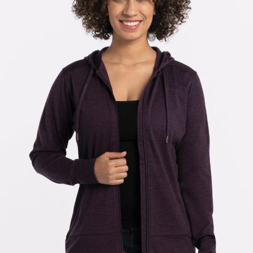 A person with curly hair is smiling, wearing the Woolx Ryann Hoodie in Deep Plum, which has a lightweight and athletic fit, over a black top and jeans. The background is plain white. | Tori is 5'7", wearing a size S