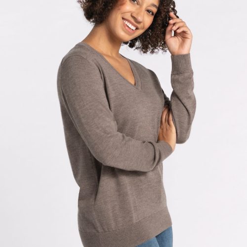 A person with curly hair smiles, wearing the Francesca Sweater in Sable from Woolx and blue jeans, standing against a plain white background. | Tori is 5'7", wearing a size S