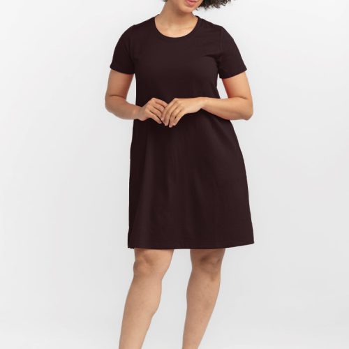 A person with curly hair stands against a white background, wearing a short-sleeved French Roast Georgie Dress by Woolx and brown wedge sandals. They have a relaxed posture, with one hand lightly touching the other. | Tori is 5'7", wearing a size S