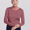 An older woman with short white hair is wearing the Woolx Remi Long Sleeve T-Shirt in Wild Ginger along with dark jeans, standing against a plain white background. She smiles gently and holds her hands together in front of her. | Kathy is 5'9", wearing a size S