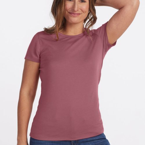 A person with long hair smiles while standing against a plain background. They are wearing the Addie Short Sleeve Crew in Wild Ginger from Woolx, made from Woolx DuraLite® Fabric, paired with blue jeans. One arm is raised, and their hand gently touches their head. | Jessie is 5'9", wearing a size S