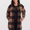 A person with long black hair is wearing the Sutton Shacket by Woolx, which features a camel checkered pattern, over a sleek black outfit. They are smiling with their hands in the shacket pockets while standing against a plain white background.