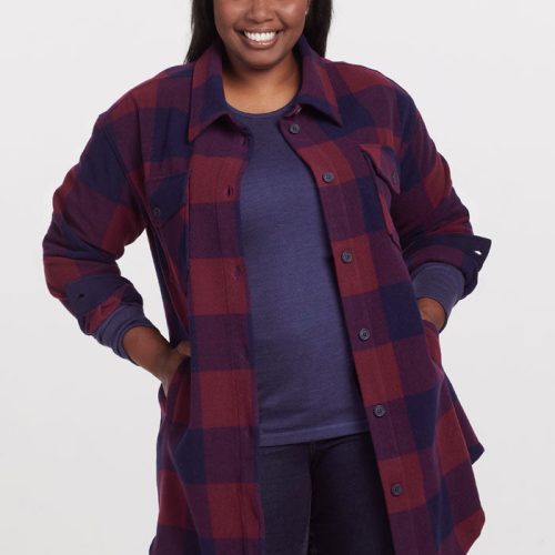 A smiling person stands against a plain background in a Sutton Shacket by Woolx—a stylish Cranberry Navy Checkered jacket layered over a blue top. Their long dark hair cascades down, and with hands comfortably tucked in their pockets, they exude effortless charm.