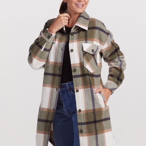 Dressed in the Sutton Shacket - Forest Plaid by Woolx, a person stands confidently with one hand on their hip and the other lightly touching their chin. The shacket features shades of green, brown, and white, offering a breathable plaid design. Complementing this look is a black top and blue jeans, all tied together with a subtle smile.