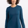 A person with curly hair is smiling in a relaxed-fit, merino wool Hazel Tunic in Real Teal by Woolx, paired with jeans. They are standing against a plain white background. | Tori is 5'7", wearing a size S