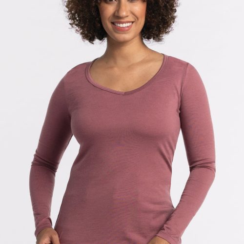 A person with curly hair smiles while standing against a plain white background. They are wearing a Wild Ginger Layla V Neck Top by Woolx and dark jeans, exuding effortless style. | Tori is 5'7", wearing a size S