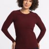 A person with curly hair is wearing the Woolx Hadley Ribbed Crew in Cranberry Melange, paired with dark jeans. They are standing against a plain white background, smiling with hands on hips, perfectly showcasing their stylish travel wardrobe. | Tori is 5'7", wearing a size S