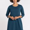 A person with curly hair is wearing the Rissa A Line Dress in Real Teal by Woolx, crafted from merino wool and showcasing three-quarter sleeves. They are standing against a plain white background, smiling, with their hands together at waist level. | Tori is 5'7", wearing a size S