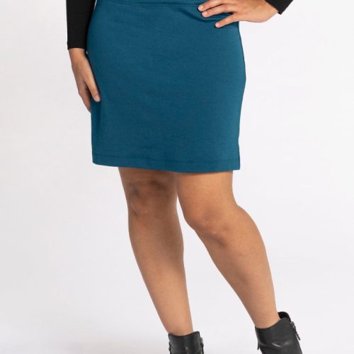 A person wearing the Woolx Monroe Knee Length Skirt in Real Teal, made from travel-friendly merino wool for its moisture-wicking properties, pairs it with a black long-sleeve top and holds one hand on their hip. They complete the look with black ankle boots against a plain white background. | Tori is 5'7", wearing a size S