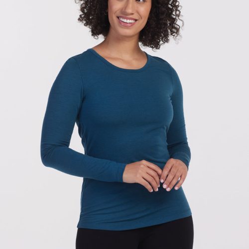 A person with curly hair wearing the Woolx Remi Long Sleeve T-Shirt in Real Teal, crafted from ultra-lightweight fabric, paired with black pants, stands smiling against a plain white background. | Tori is 5'7", wearing a size S