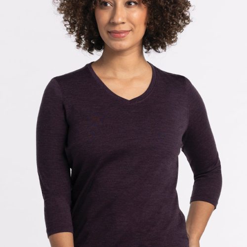 A person with curly hair is smiling and striking a pose with one hand on their hip. They are wearing a relaxed fit Elena V Neck in Deep Plum by Woolx, paired with jeans, against a plain light gray background.