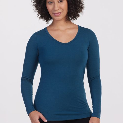 A person with curly hair is dressed in the fitted, long-sleeve Layla V Neck Top in Real Teal by Woolx and black pants, standing against a plain white background, smiling gently. | Tori is 5'7", wearing a size S