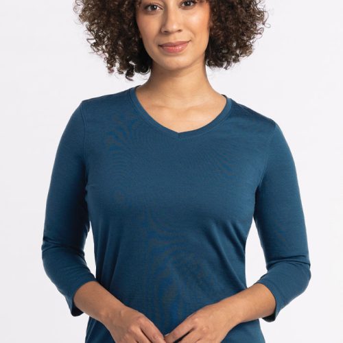 A person with curly hair is wearing a Real Teal Elena V Neck by Woolx, known for its relaxed fit. They are standing against a plain white background. | Tori is 5'7", wearing a size S