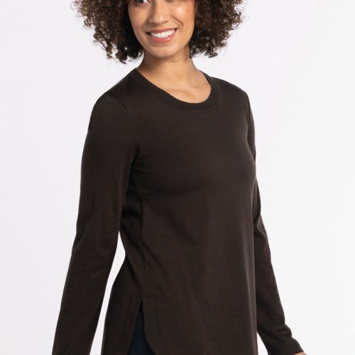 A person with curly hair smiles while wearing the Woolx Hazel Tunic in French Roast, a long-sleeved top made of lightweight fabric. The tunic features a round neckline and side slits as they stand against a plain white background. | Tori is 5'7", wearing a size S