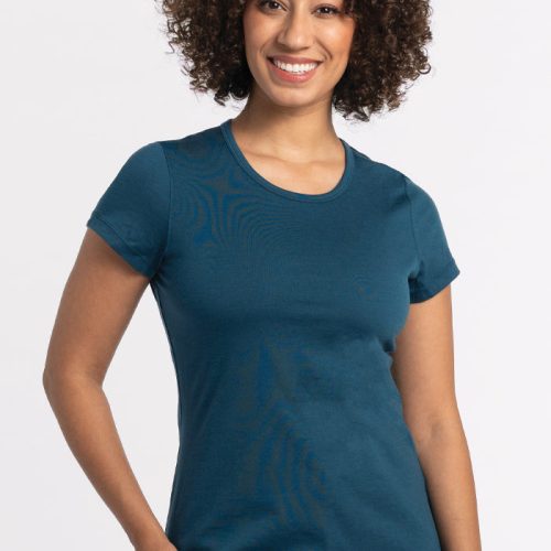 A person with curly hair is smiling while wearing a Woolx Addie Short Sleeve Crew in Real Teal, an odor-free tee crafted from Australian Merino Wool. They are standing against a plain white background. | Tori is 5'7", wearing a size S