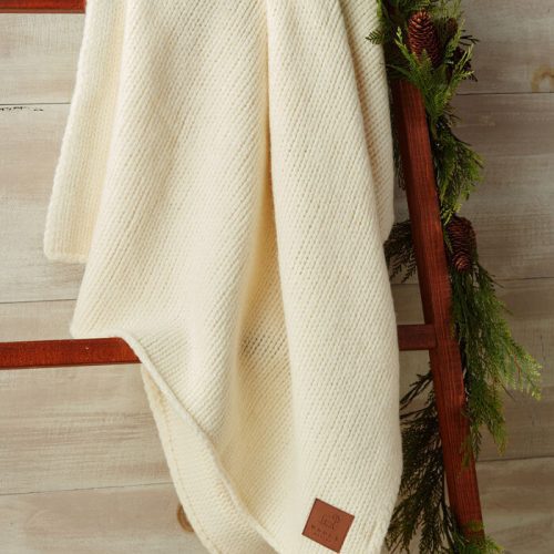 Draped over a wooden ladder is *The Saranac Throw* by Woolx, a heavyweight cream-colored blanket featuring an intricate textured pattern. The ladder is adorned with festive evergreen branches and pinecones, set against a rustic wood backdrop. A small leather tag highlights the blanket's Woolmark Certification.