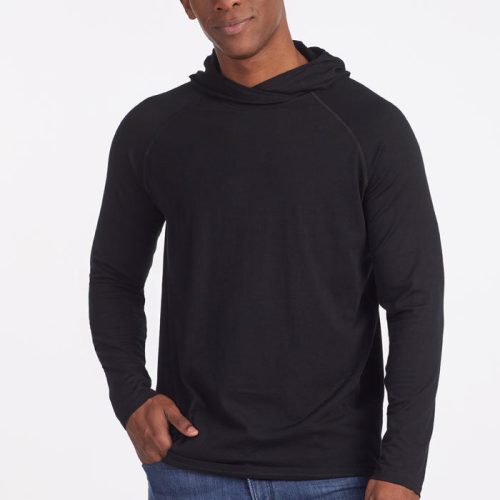 A person dressed in a black "Base Camp Hoodie OLD" from Woolx, paired with blue jeans, stands against a simple white background with hands in pockets and a slight smile.