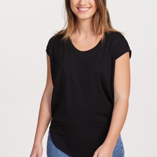 A person with long hair, wearing a Gabi Tunic Top by Woolx in black and blue jeans made of lightweight fabric, smiles at the camera. The background is plain and light-colored.