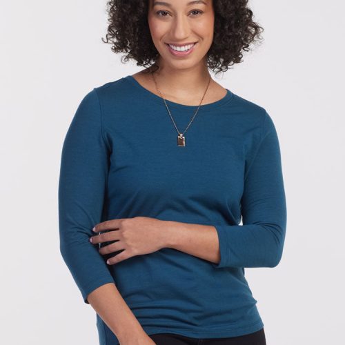 A person with curly hair smiles at the camera, adorned in a Real Teal Jenny 3/4 Sleeve Crew Neck by Woolx and a necklace. They stand against a plain white backdrop, effortlessly stylish in their lightweight top. | Tori is 5'7", wearing a size S