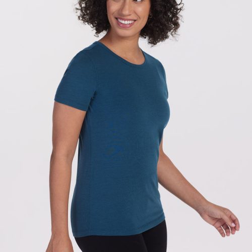 A person with curly hair, dressed in the Woolx Liza Crew Neck Tee in Real Teal and black pants, is smiling and looking to the side against a plain white background. | Tori is 5'7", wearing a size S