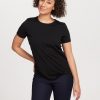 A person with curly hair is wearing a Woolx Liza Crew Neck Tee OLD in black, crafted from moisture-wicking fabric, paired with dark jeans. They are standing against a plain background, leaning slightly to one side and displaying a gentle smile.