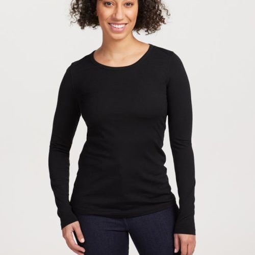 A person with curly hair is smiling and posing in a Woolx Remi Long Sleeve T-Shirt OLD in black, paired with dark pants against a plain backdrop. | Tori is 5'7", wearing a size S