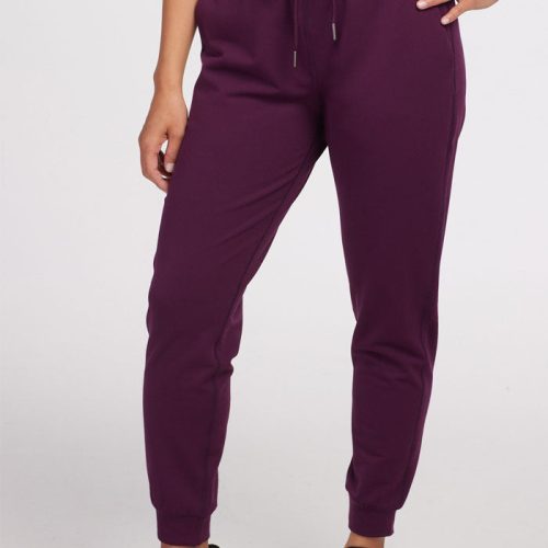 A person wearing the Woolx Lola Jogger in Winter Bloom, crafted from recycled polyester, stands against a plain background. The plum-colored joggers, showcasing a drawstring waist, are paired with black sneakers. With their midsection and legs visible, their arms rest casually at their sides.