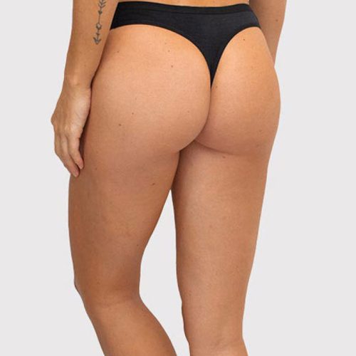 Rear view of a person wearing a black Kylie Thong Underwear by Woolx paired with a matching top. A small tattoo is visible on the left arm. The lightweight design offers no-show panty lines, set against the plain, light-colored background.