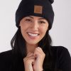 A woman with long, dark hair is smiling and looking directly at the camera. She is wearing a Woolx Baylor Beanie in Carbon Black, adorned with a brown patch featuring an animal logo. Dressed in a black top, she has her hands clasped near her face against a plain white background.